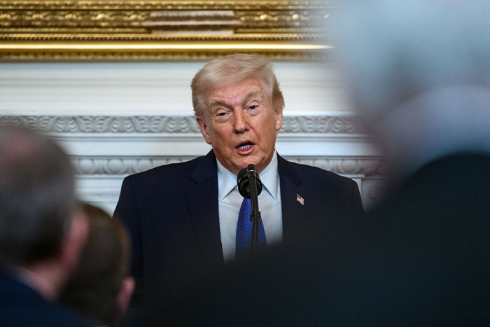 Donald Trump speaking at a podium in a formal setting with audience members visible in the foreground.