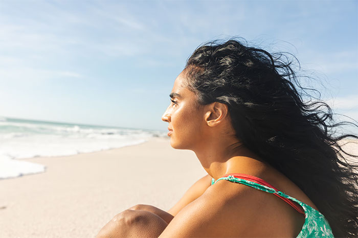 Woman with long hair sitting on beach looking at ocean, reflecting after online argument with husband about being right.