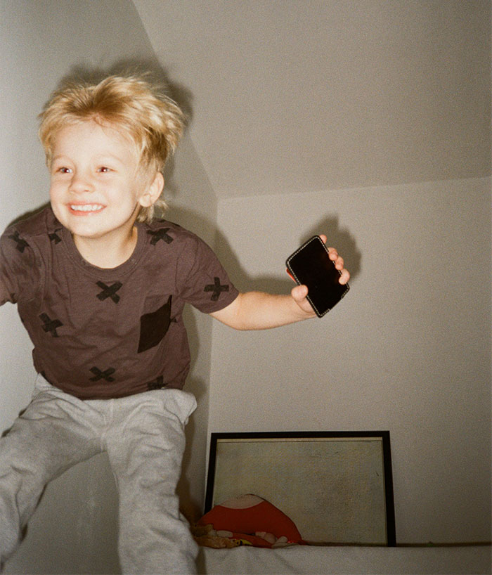 Smiling young boy holding a phone, jumping indoors, illustrating sibling and parenting challenges in a family setting.