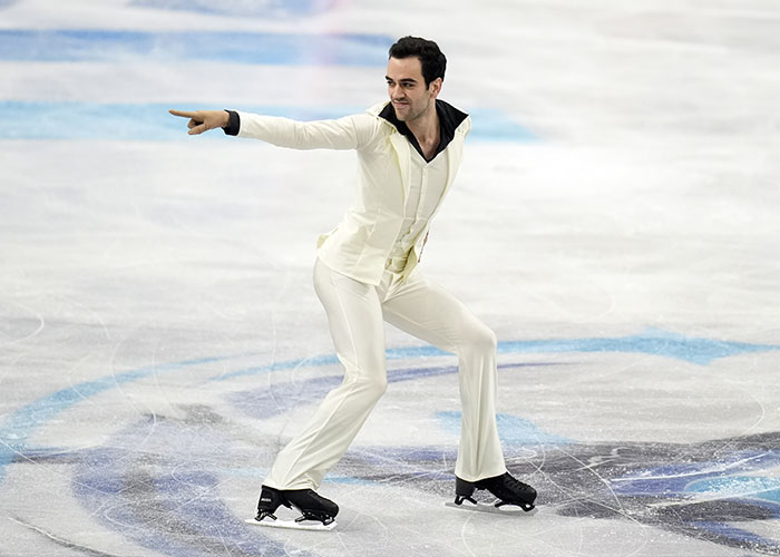 Male Olympic figure skater performing on ice rink wearing cream outfit during Winter Games competition.