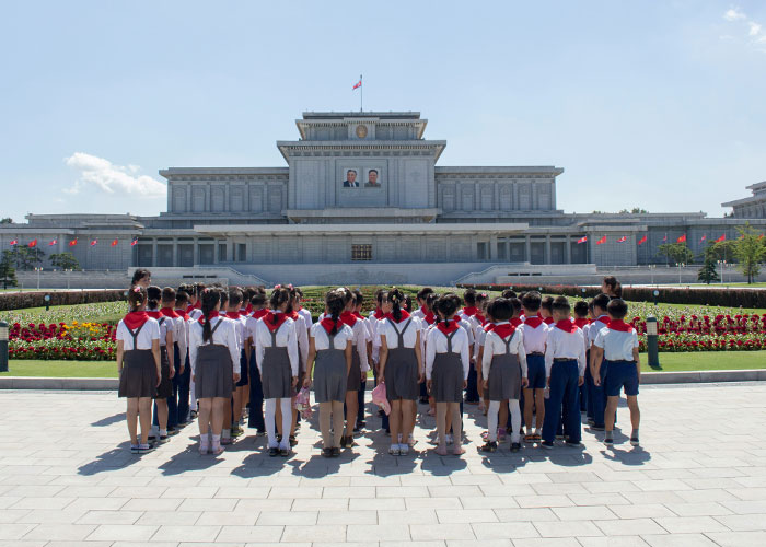 Group of North Korean children in uniform facing a government building with portraits and national flags on a clear day.