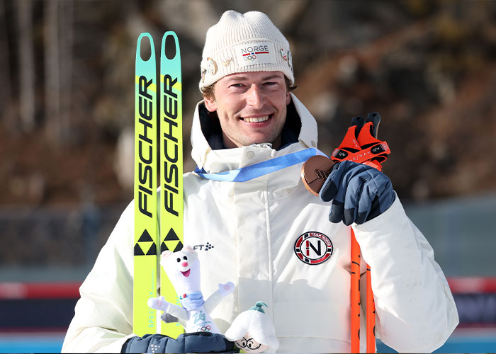 Olympic medalist holding skis and bronze medal, smiling with winter gear after medal victory in outdoor setting.