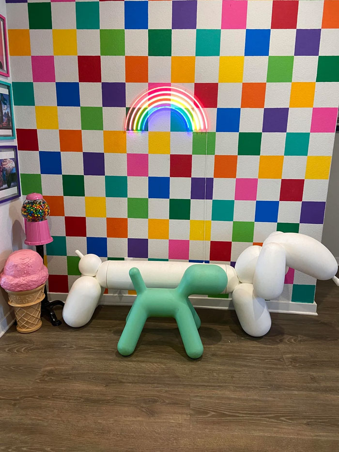Colorful checkered wall with neon rainbow light, balloon dog sculptures, and oversized candy decor in a vibrant eclectic home setting.