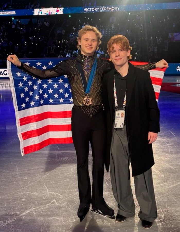 Ilia Malinin on ice holding American flag with medal after Olympic performance alongside a companion at victory ceremony.