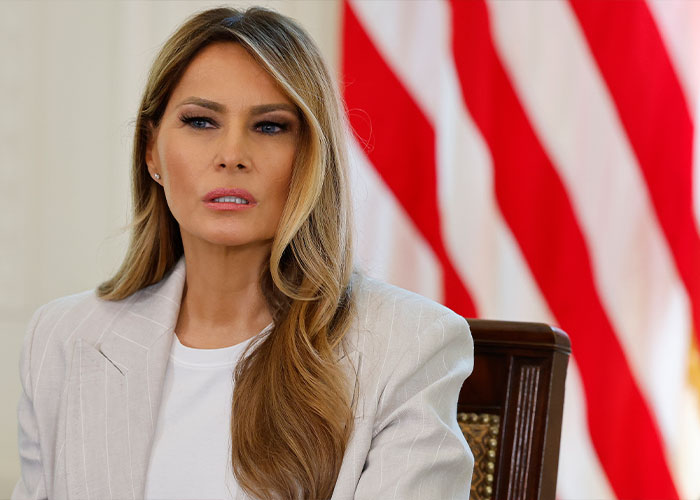 Melania Trump seated in a white blazer with American flags in the background during a formal event.