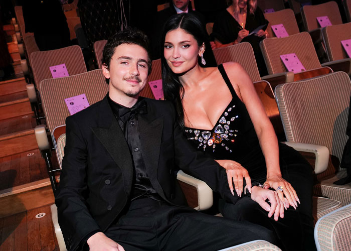 Kylie Jenner wearing a black dress with embellishments, seated next to a man in a black tuxedo at the 2026 BAFTAs event.