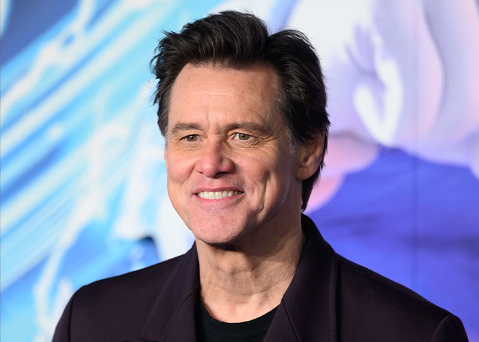 Jim Carrey smiling at a public event, wearing a dark jacket with a blue and purple blurred background.