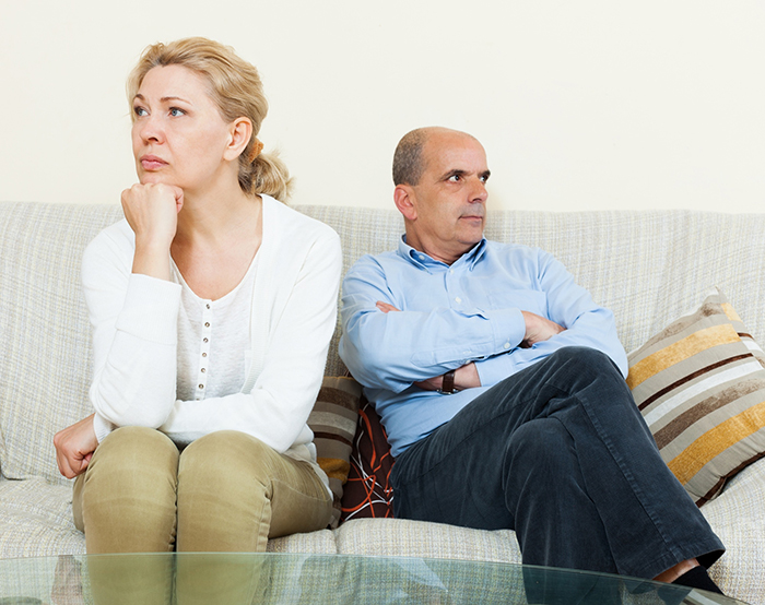 Middle-aged man and woman sitting apart on a couch, appearing upset and refusing to communicate, illustrating family conflict.