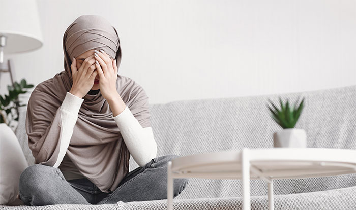 Woman wearing hijab sits on couch covering face, expressing distress weeks after marrying husband and exposing his secret.