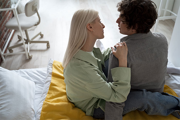 Couple in serious conversation on bed, highlighting tension after family needed to move in with them.