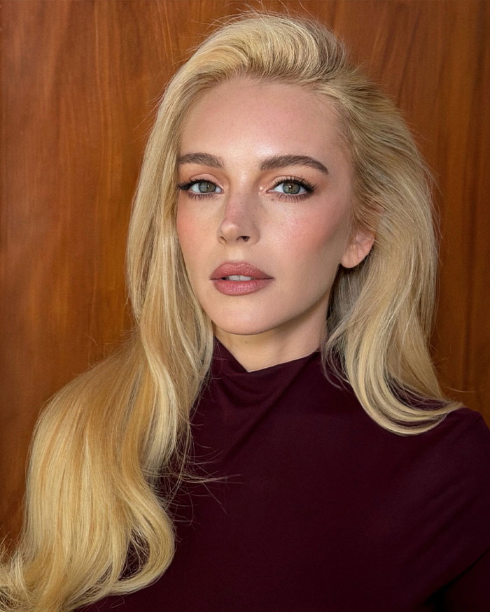 Lindsay Lohan with long blonde hair and natural makeup posing against a wooden background in a maroon top.