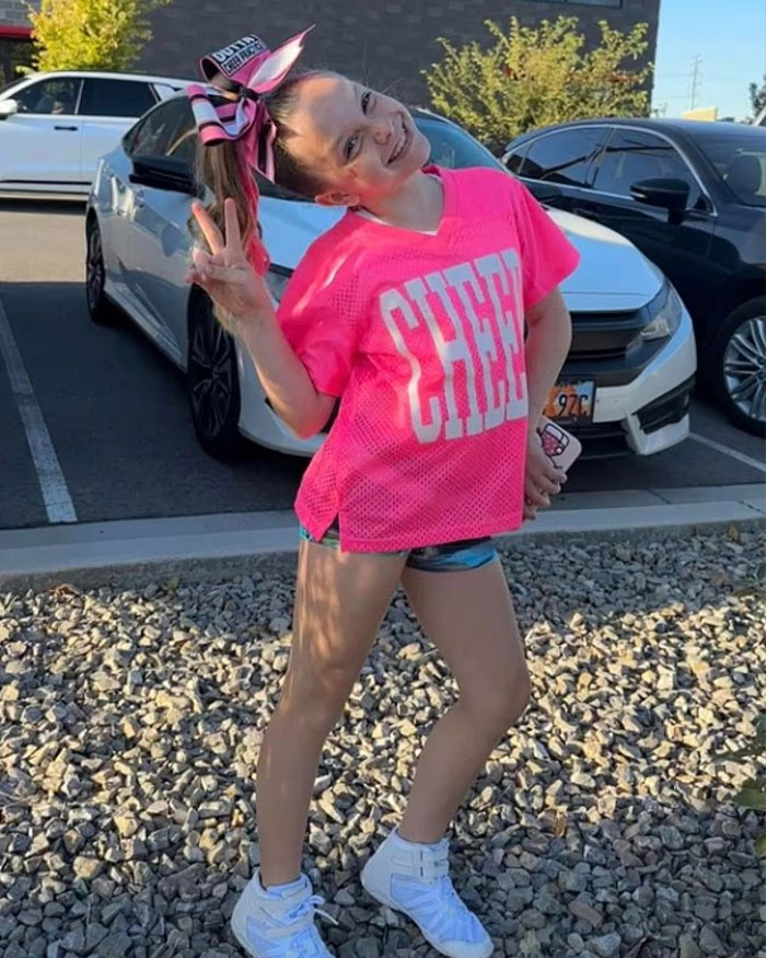 Young girl in bright pink cheer outfit posing outdoors with a peace sign, related to Utah Dance Mom&rsquo;s mean texts and parent clash.