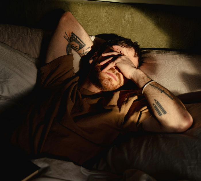 Man waking up in bed covering his face, showing tiredness in a dimly lit room, depicting normal couples waking each other up.