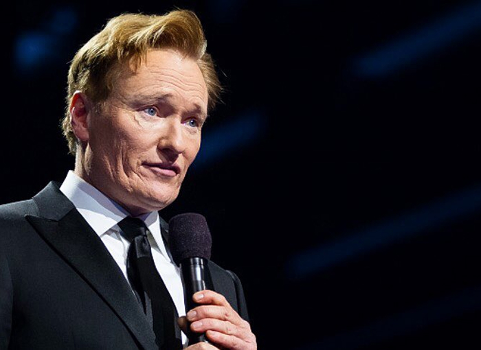 Conan O&rsquo;Brien speaking on stage holding a microphone during a formal event about Oscars hosting return.