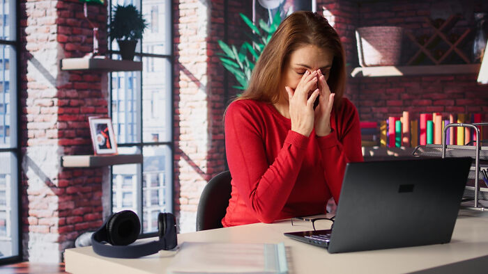 Stressed woman in red sweater rubbing eyes in office, experiencing toxic workplace moments and frustration at her desk.