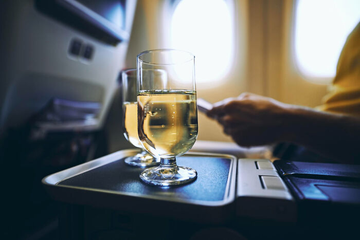 Two glasses of white wine on an airplane tray table with a passenger using a phone, highlighting underrated plane perks.