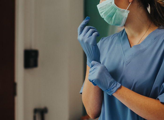 Healthcare worker wearing scrubs and mask putting on gloves, representing wife working longer shifts in a demanding job.