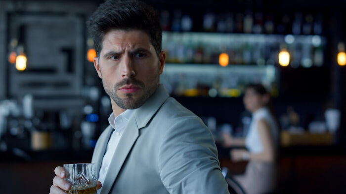 Man in a light gray suit holding a drink at a bar, looking surprised during a wild dating experience.