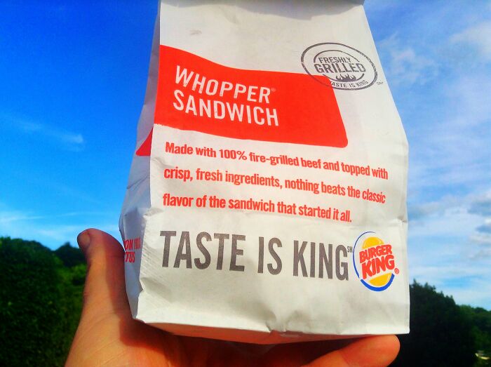 Hand holding a Burger King Whopper sandwich bag against a blue sky and green trees background.