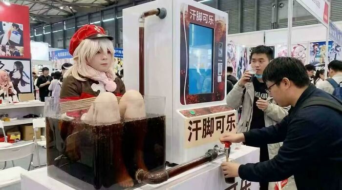 Person in cosplay sitting in a giant soda machine filled with dark liquid while others watch and pour drinks at an event.