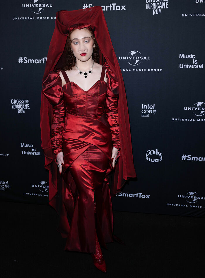 Chappell Roan at a Universal Music event wearing a bold red satin gown with a dramatic headpiece and necklace.
