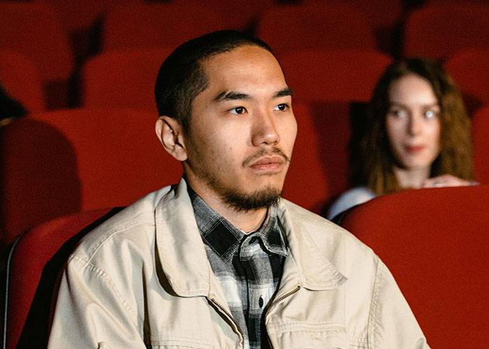 Young man in a beige jacket sitting in a theater, reflecting on today I messed up stories with a woman blurred in the background.