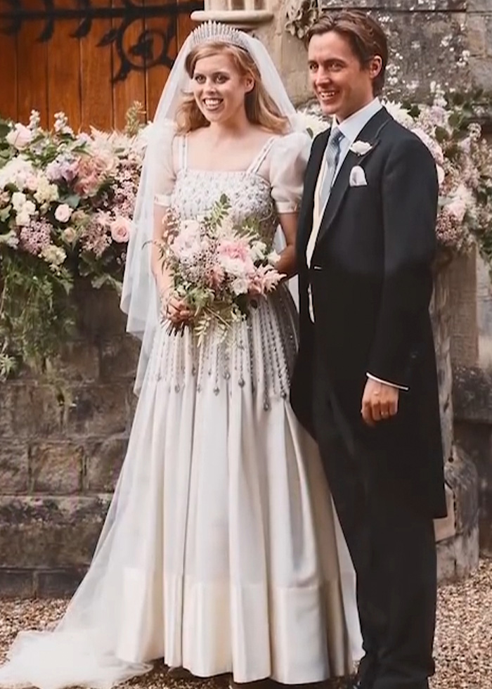 Bride in a vintage-style royal wedding dress holding a bouquet, standing next to groom in formal attire outdoors.