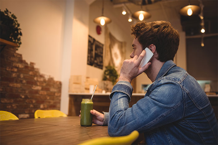 Young man on a first date talking on phone, holding a green smoothie, in a cozy cafe setting.