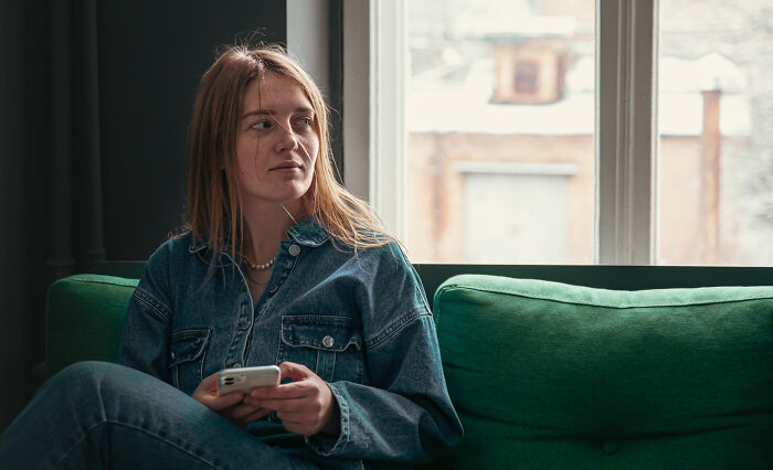 Young woman in denim outfit holding phone, sitting on green couch, reflecting on wild dating experiences that made her question reality.