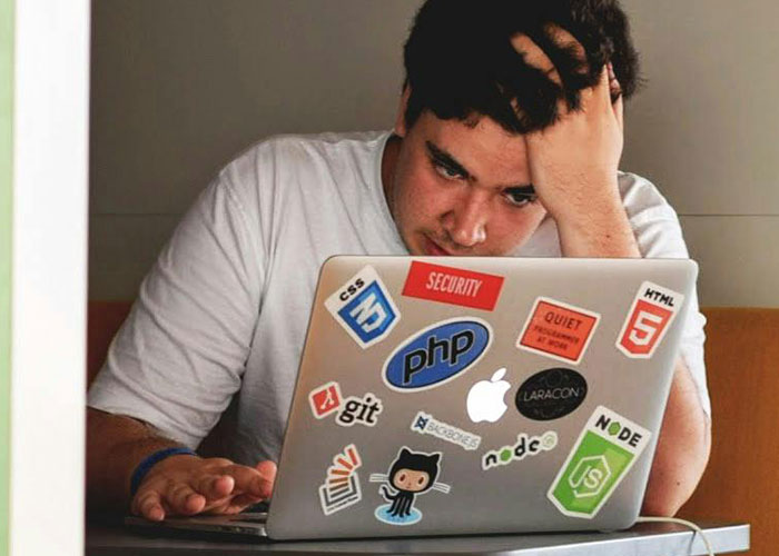 Man looking stressed while working on laptop covered with programming and security stickers, reflecting messed up stories.