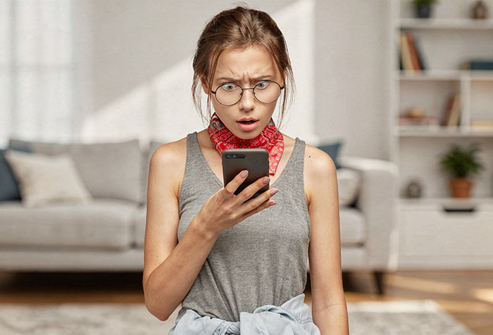 Young woman wearing glasses and a bandana looks shocked while holding a phone, representing K-Pop fan merch dispute.