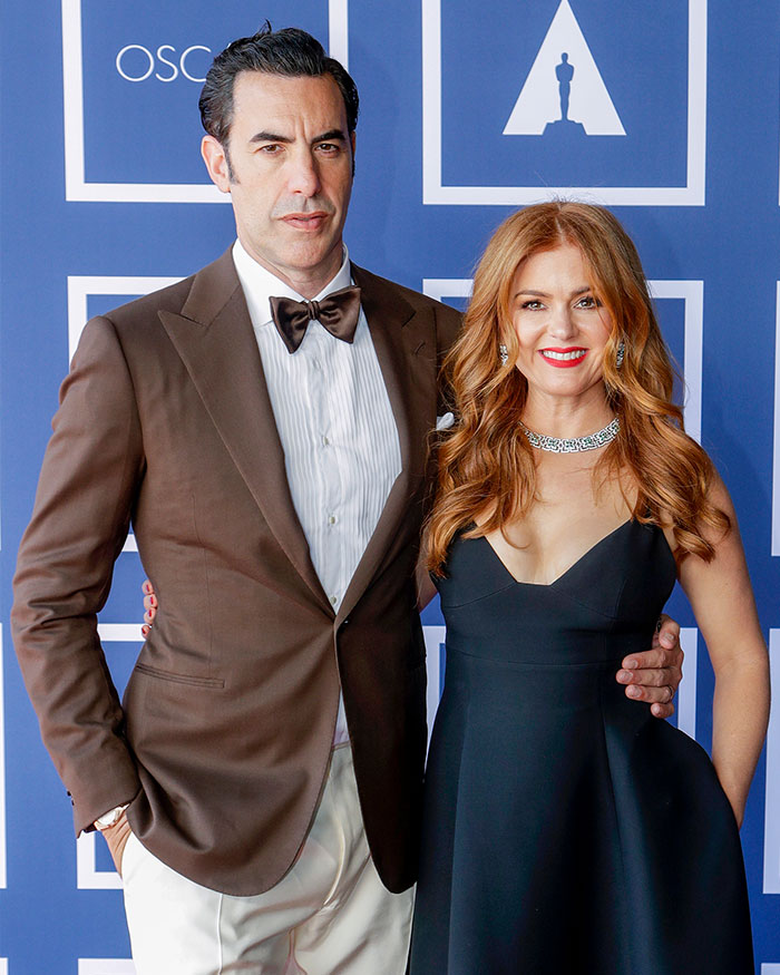 Sacha Baron Cohen and Isla Fisher posing together at an event, dressed formally in tuxedo and black dress.