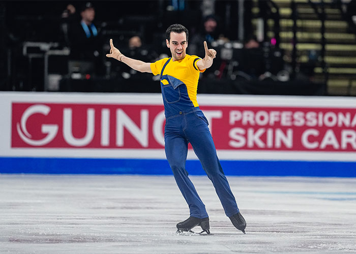 Male Olympic figure skater in a yellow shirt and blue overalls performing on ice with a unique music choice at Winter Games.