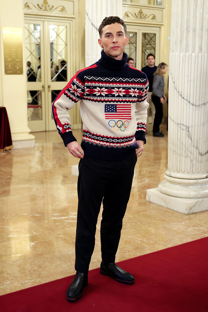Man wearing a festive Olympic sweater and black pants posing indoors on a red carpet at Olympics 2026 event.