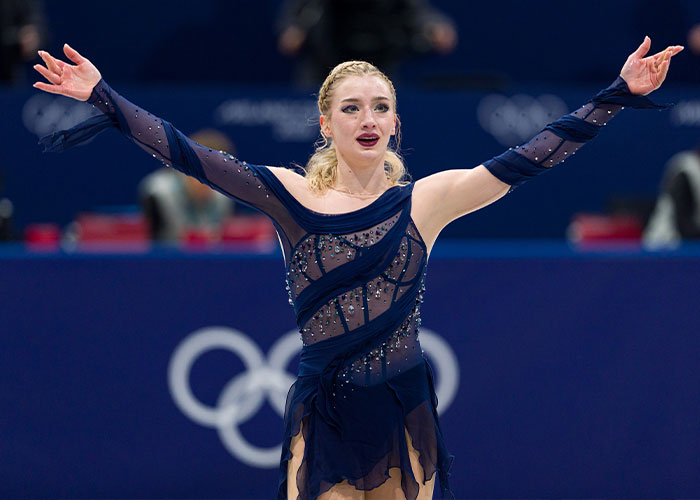 Amber Glenn on ice in a navy blue dress, performing with arms extended at a major skating competition.