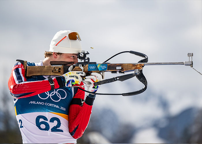 Olympic medalist aiming rifle during winter biathlon event at Milano Cortina 2026 with snow-covered mountains in background.