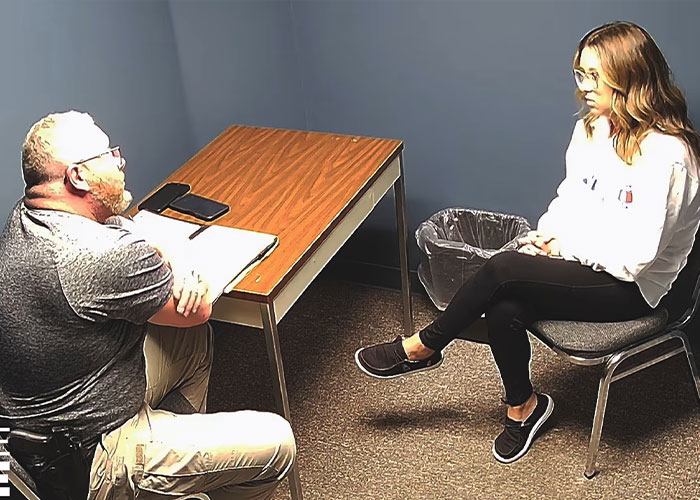 School employee breaking down during confession while sitting opposite a man in a small room with a wooden table.