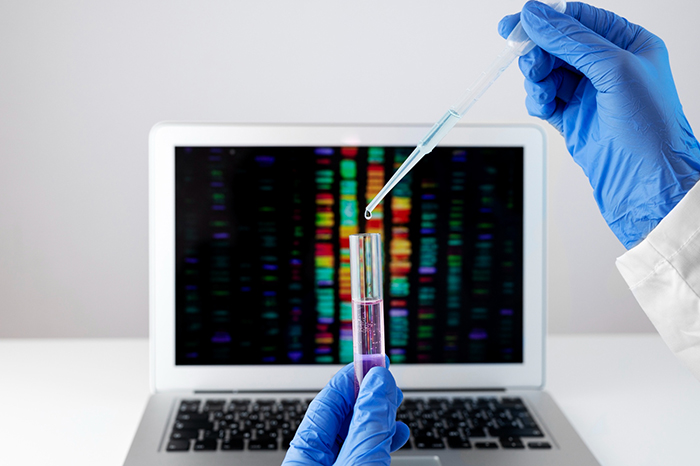 Scientist wearing blue gloves holding a test tube and dropper with DNA sequence analysis on laptop screen in background.
