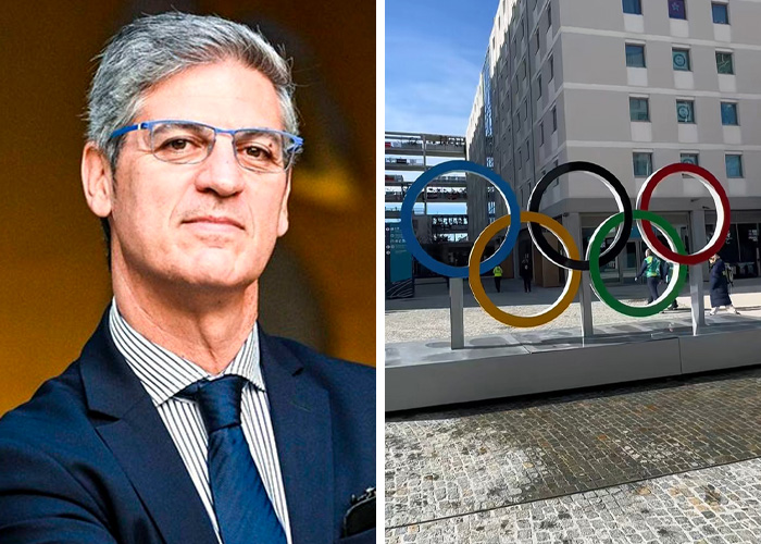 Man in a suit and glasses next to large Olympic rings sculpture at outdoor 2026 Winter Olympics venue.