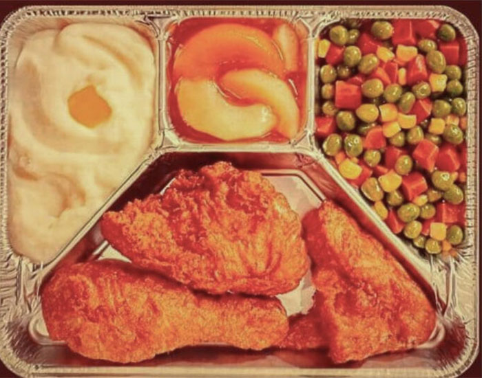 Nostalgic TV dinner tray with fried chicken, mashed potatoes, mixed vegetables, and fruit cocktail in foil container.