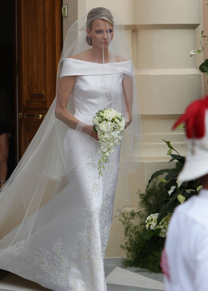 Bride in an elegant royal wedding dress with off-shoulder neckline and delicate floral embroidery holding a cascading bouquet.