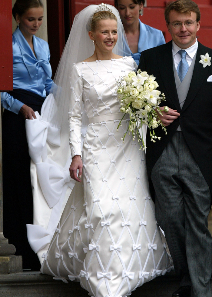 Bride in a long-sleeve royal wedding dress with bow details and a tiara, holding a white floral bouquet walking with groom.