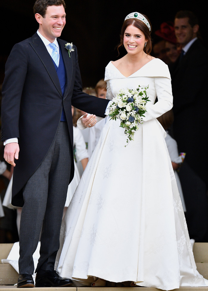 Bride in a royal wedding dress holding a bouquet, standing next to groom in formal attire, smiling outdoors.