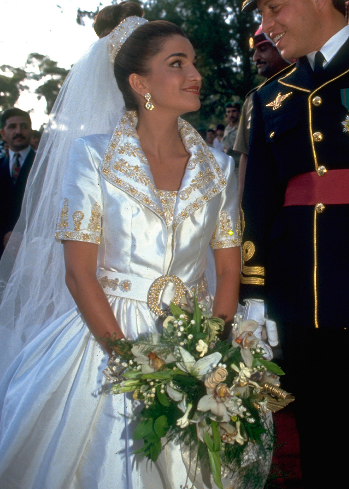 Bride in a royal wedding dress with gold embroidery holding a bouquet, standing next to a man in formal military attire.