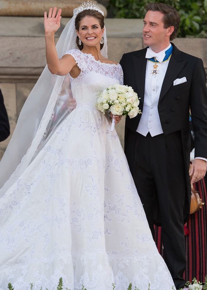 Bride wearing a lace royal wedding dress holding a bouquet, standing next to groom in formal attire, waving and smiling.