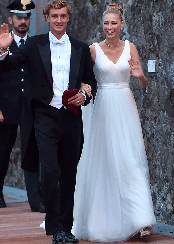 Bride and groom walking outdoors, bride wearing elegant white royal wedding dress, both smiling and waving.