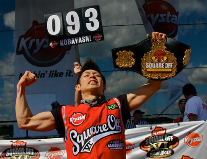Competitive eater Kobayashi celebrating with a championship belt and scorecard, showcasing record-setting facts in contests.