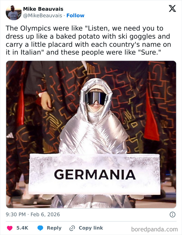 A person in a silver padded outfit and ski goggles holding a sign labeled Germania during the 2026 Winter Olympics.