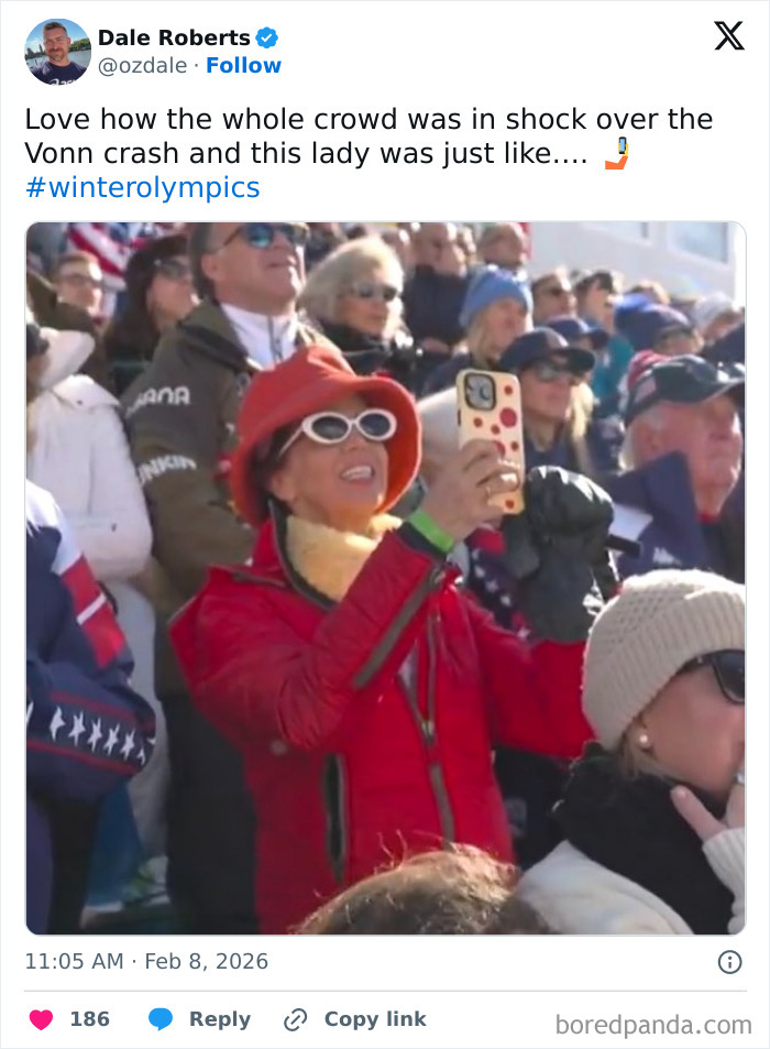 Woman in red jacket and hat taking a photo of Winter Olympics crowd reacting to incident during 2026 event.