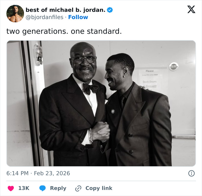 Michael B. Jordan reacting with a smile, dressed in a suit, sharing a moment with an older man in formal attire.
